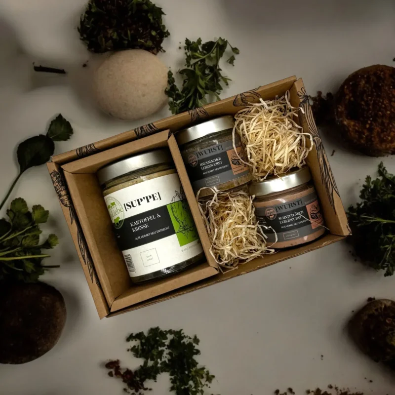 Geschenkbox mit Kartoffel-Kresse-Suppe und zwei Gläser Leberwurst, umgeben von Kräutern