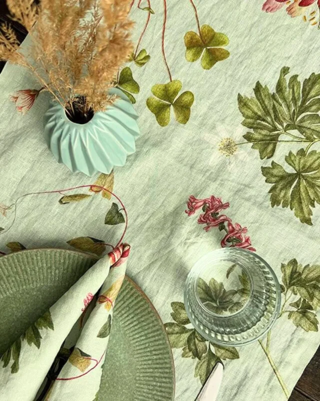 Der Leinen-Tischläufer "Flora Danica Atlas" mit botanischen Motiven, dekoriert mit einer hellgrünen Vase, getrockneten Gräsern und Blättern. Die natürliche Optik des Leinens passt perfekt zur ländlichen Tischdekoration.
