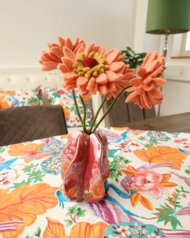 Blumenvase "Idrija", "Gerbera" Filzblumen und eine "Pai" Mitteldecke stehen auf einem Holztisch im Esszimmer. Die Mitteldecke ist farbenfroh mit abstrakten Blumen und Kranichen gestaltet und im Hintergrund sind auf dem Sofa die passenden Kissen zu sehen. Die Blumenvase ist knallig Rot und mit Filzblumen gefüllt.