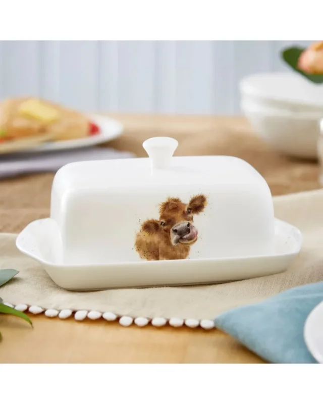 Kuh Butterdose "Royal Worchester" aus Porzellan von Wrendale Designs auf einem bedeckten Tisch. Das Motiv zeigt eine Kuh mit herausgestreckter Zunge. Im Hintergrund sind Teller zu sehen.