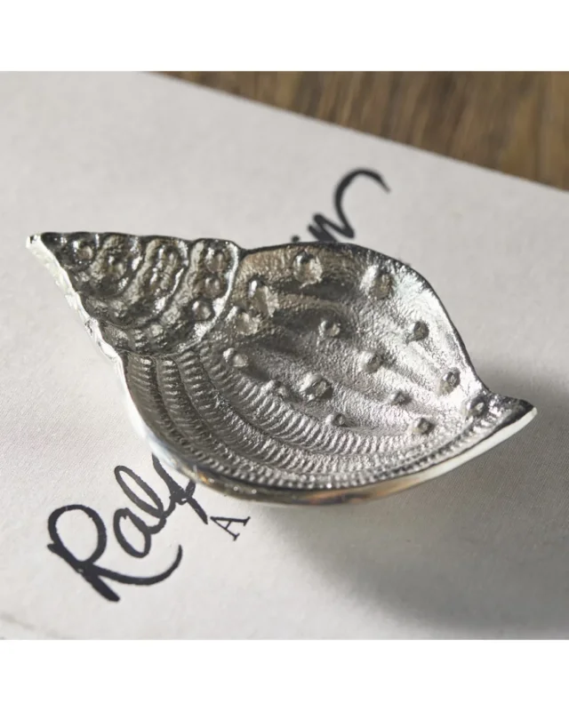 Silberne dekorative Muschelschale von Rivièra Maison auf einem weißen Blatt welches auf einem hellen Holztisch liegt.