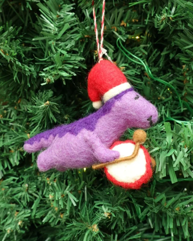 Dekorierter lila "Dino mit Trommel" Weihnachts-Filzanhänger von Grace & Gloria hängt an einem Tannenbaum. Der Dino ist Dunkel und Helllila. Er trägt eine rote Weihnachtsmütze und eine Trommel.