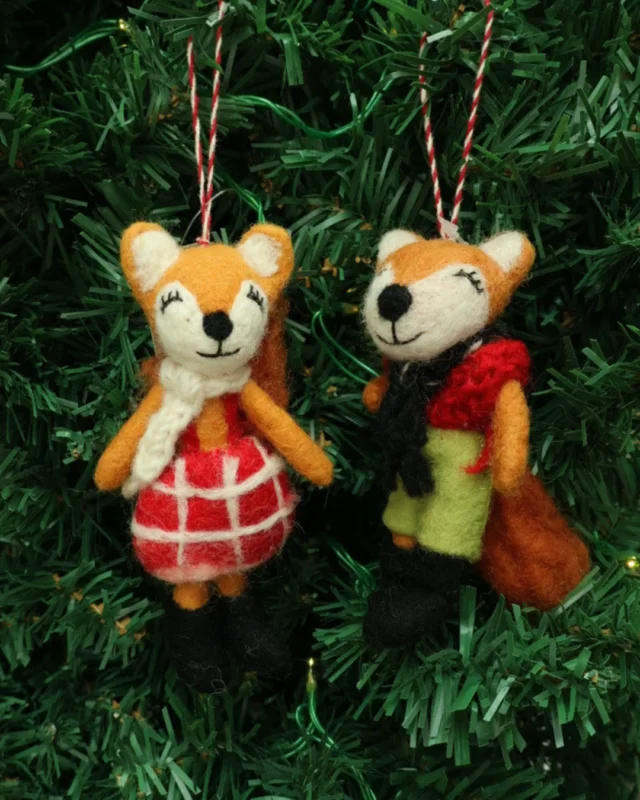 Dekorierter Weihnachts-Fuchs-Filzanhänger "Naomi und Nils" von Grace & Gloria hängend an einem Tannenbaum. Es sind 2 süße Füchse mit einer grünen Hose und einem roten Kleid.