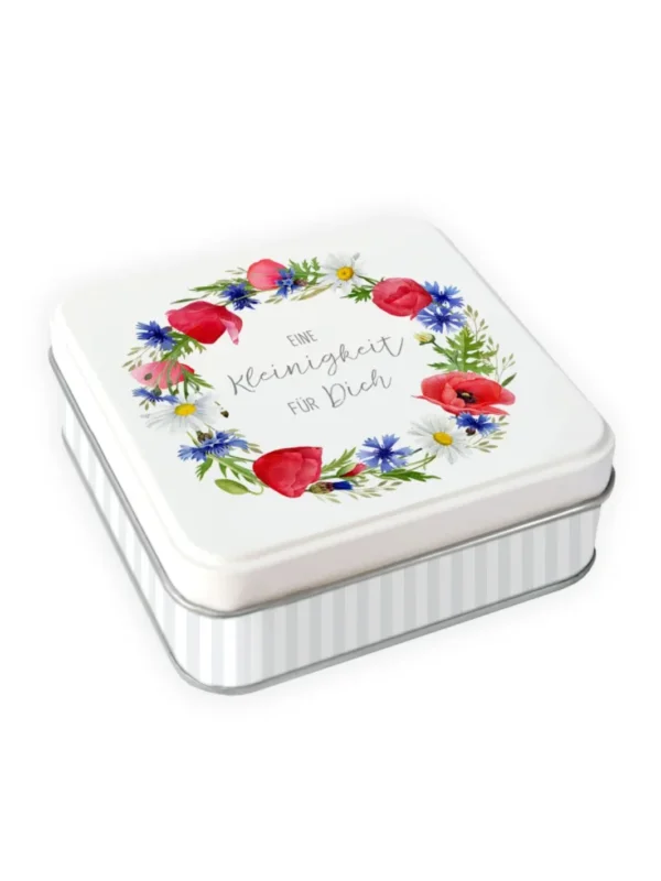 Quadratische Geschenkdose vom Grätz Verlag mit weißem Deckel, floralem Blumenkranz aus roten, blauen und weißen Blüten und Schriftzug „Eine Kleinigkeit für Dich“, hell gestreifter Unterteil, freigestellt vor weißem Hintergrund, leicht schräg fotografiert.