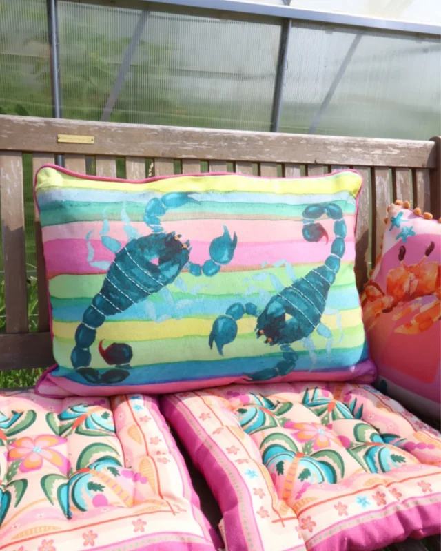 Buntes sommer outdoor Kissen "Skorpion". Das abstrakte Motiv besteht aus 2 Türkisen Skorpionen und einem bunt gestreiften Hintergrund. Das Kissen liegt auf einer farblich passend dekorierten Bank.