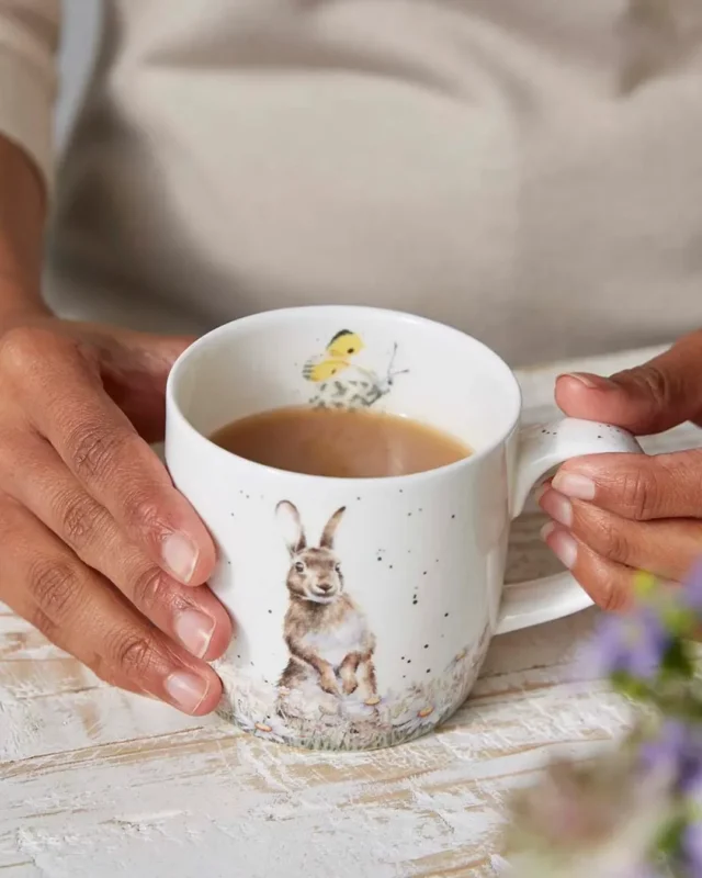 Hasen Tasse von Wrendale Designs mit Kaffe gefüllt auf hellen Holztisch. Auf der Tassen Außenseite ist ein süßer Hase in einer Blumenwiese zu sehen. Auf der Innenseite ist ein gelber Schmetterling.