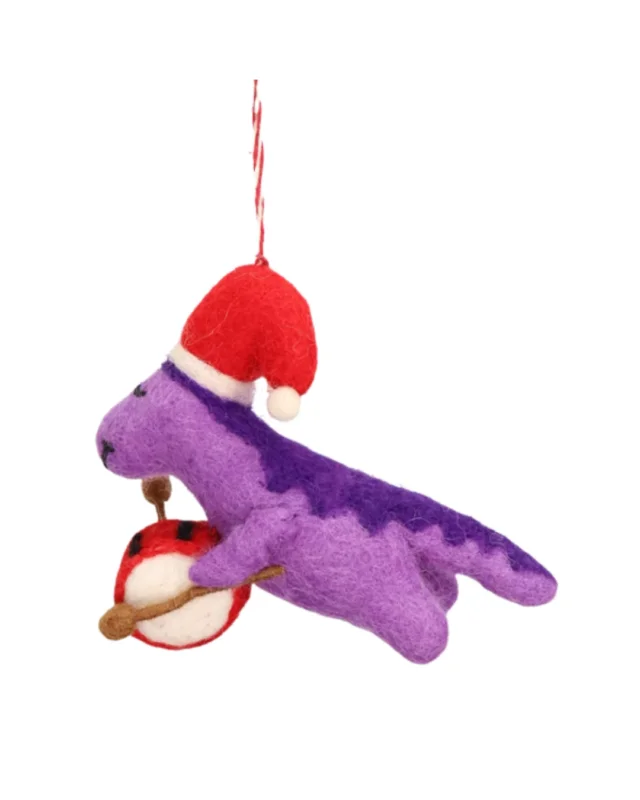 Seitansicht des lilafarbenen Weihnachtsdino Filzanhängers von Grace & Gloria. Der freundlich schauende Dino trägt eine Rote Weihnachtsmütze auf dem Kopf und eine rot-weiße Trommel vorm Bauch, welche er mit zwei dazugehörigen Trommelstöcken spielt.