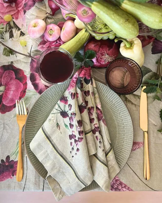 Zwei Leinenservietten Blossom Garden von Koustrup & Co., dekorativ gefaltet auf einem grünen Teller, kombiniert mit goldfarbenem Besteck.
