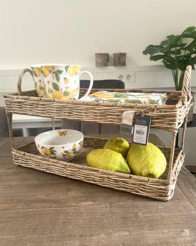 Zitronen Geschirr "Fresh Lemons" von Ambiente und Riviera Maison auf einem Holztisch. Das Rattan Etagere ist von Riviera Maison die 2 Tassen, Schüssel und die Servietten sind von Ambiente. Im Hintergrund sind eine Zimmerpflanze und 2 Stühle zu sehen.
